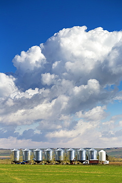 A row of large metal grain bins with dramatic storm clouds and blue sky in the background, West of Calgary, Alberta, Canada