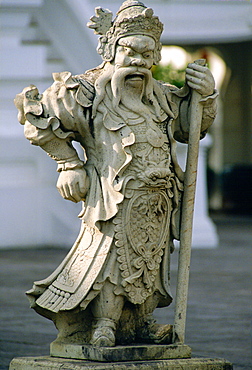 An ancient stone statue of a man dressed in long robes holding a staff outside the Grand Palace, Bangkok, Thailand.