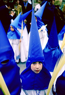 Young girl staring out from her blue silk hooded costume while taking part in the Semana Santa parade for Holy Week in Seville, Spain (Sevilla). They are carrying palm leaves.