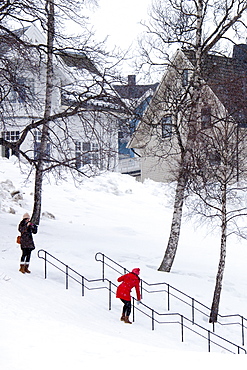 Young woman takes photographs as her friend hangs onto handrail to avoid slipping on the steps in snowy Tromso, Norway