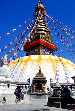 Children sitting on the wall outside theSwayambhunath Stupa  in Kathmandu valley in Nepal which is bedecked with flags.  It is a sacred monument in the Buddhist world and is a symbolic representation of the fully enlightened mind and the path to enlightenment.