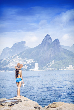 A 20-25 year old young Brazilian woman standing on the Arpoador rocks with Ipanema and the Morro Dois Irmaos hills in the distance, Rio de Janeiro, Brazil, South America