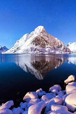 The snowy peaks are reflected in the frozen sea on a starry winter night, Reine Bay, Nordland, Lofoten Islands, Arctic, Norway, Scandinavia, Europe