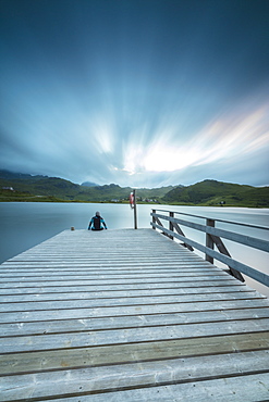 Hiker on wooden deck admires the sea illuminated by blue lights at night, Holdalsvatnet, Vestvagoy, Lofoten Islands, Norway, Scandinavia, Europe