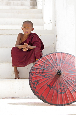 Young monk dressed in red, sits on steps with red parasol at the Myatheindan Pagoda (White Temple) in Mingun, Myanmar (Burma), Asia