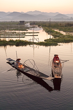 Two fishermen keep warm in their long tail fishing boats at dawn near the floating gardens on Inle Lake, Shan State. Myanmar (Burma), Asia