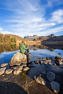 Walker wearing green jacket sat admiring the mirror conditions at Blea Tarn, Lake District, Cumbria
