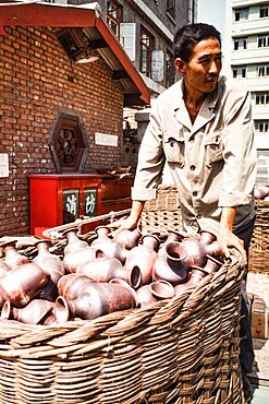 A worker in a cloisonne workshop with a basket of bronze vases to finished as cloisonne. Beijing, China.