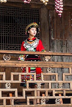 An attractive young woman in traditional ethnic Tujia dress poses for a portrait in Furong, China. Bundles of colored onions hang from the eaves as decoration.