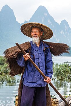 A cormorant fisherman in traditional cape on the Li River. Xingping, China, with limestone karsts behind.