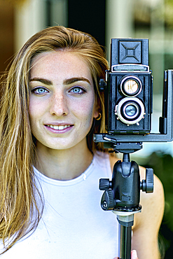 Portrait of a young beautiful caucasian woman in her 20´s with blue eyes photographing with an old vintage camera on a tripod outdoor. Lifestyle concept.