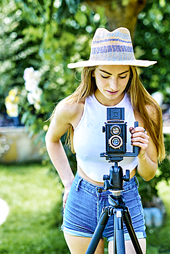 Portrait of a young beautiful caucasian woman in her 20´s with blue eyes wearing a hat and photographing with an old vintage camera on a tripod outdoor in a garden. Lifestyle concept.