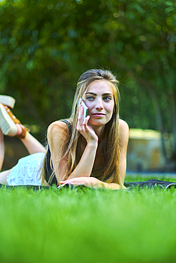 Portrait of a young beautiful caucasian woman in her 20s lying on the grass and talking on her mobile phone in a garden. Lifestyle concept.