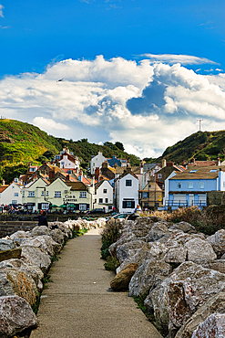 Pathway leading to a quaint coastal village with white houses, nestled between green hills under a blue sky with clouds in Staithes, England.