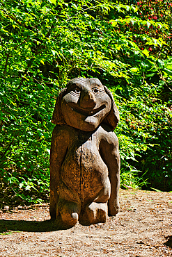 A wooden sculpture of a smiling cartoon-like character with a large head and short body, placed outdoors in a forested area.