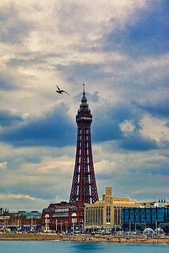 A scenic view of a tall tower, possibly a landmark, against a cloudy sky. The foreground features a beach with people and buildings, while a bird flies in the sky.