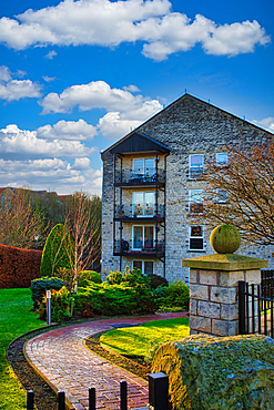 Three-story stone apartment building with balconies, landscaped grounds, and brick pathway. Sunny day with partly cloudy blue sky in Wetherby, UK.