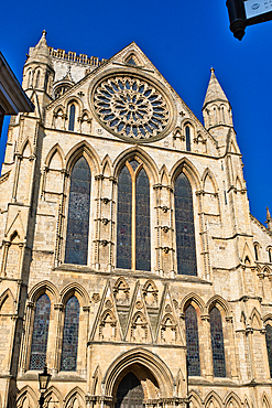 A detailed view of a beige stone cathedral facade with large stained glass windows, intricate arches, and a prominent rose window against a blue sky in York, UK.