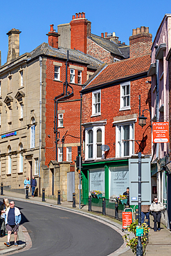 Street scene featuring brick buildings, people walking, and commercial storefronts under a bright, clear sky in Ripon, UK.