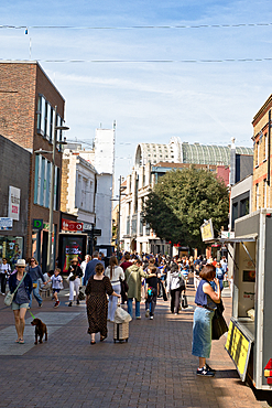Crowded pedestrian street scene with diverse buildings, a food truck, and many people walking on a sunny day in Kingston, UK.