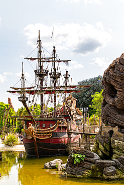 A large, decorative pirate ship is docked in a green pond with detailed rock formations and lush green foliage at Disneyland Paris.
