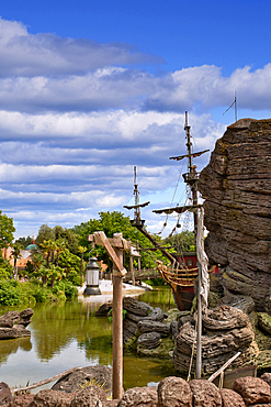 Scenic view of a pirate ship docked in a lagoon, surrounded by rocks, greenery, and a partly cloudy sky at Disneyland Paris, France.