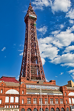 The Blackpool Tower is shown from a low angle, dwarfing the brick structure beneath. Blue sky and white clouds provide a contrasting backdrop.