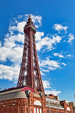 A low-angle view of a tall, red latticework tower on top of a red brick building, set against a blue sky dotted with white clouds.