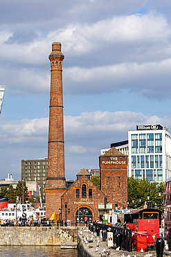 A brick smokestack tower stands tall beside a building complex. Modern structures and a body of water are visible in the background in Liverpool, UK.