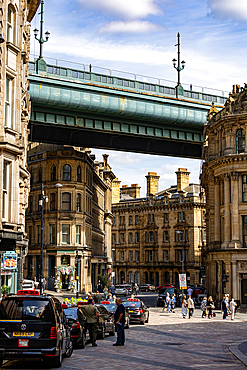 A view through city buildings towards a large, green bridge spanning over a street with taxis and pedestrians in Newcastle upon Tyne, UK.