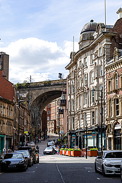 A sunlit street in a city, featuring a large stone railway arch, ornate buildings, and cars. The sky has scattered clouds in Newcastle upon Tyne, UK.