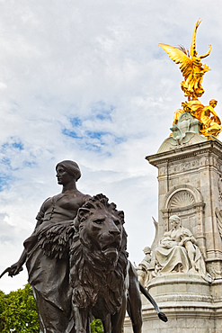 A bronze statue of a woman with a lion stands before a tall, ornate monument topped with a gold figure against a cloudy sky in London, UK.