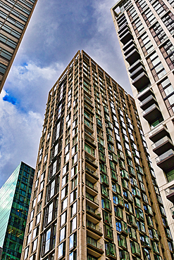 Low-angle view of several tall buildings, including a beige, glass-windowed high-rise and a teal, glass-faceted building, against a cloudy sky in London, UK.