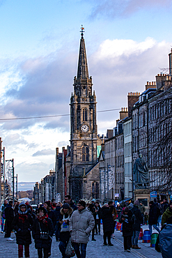 A crowded street with a tall, stone clock tower dominating the background. People are walking in both directions, dressed in winter clothing in Edinburgh, Scotland.