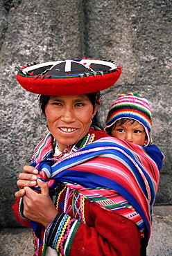 Portrait of a local woman in traditional dress, carrying her baby on her back, in front of Inca ruins, near Cuzco, Peru, South America