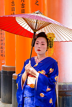 Portrait of a geisha holding an ornate red umbrella in front of a line of red torii gates, Fushimi-Inari Taisha, Kyoto, Kansai Region, Honshu, Japan, Asia