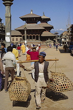 Nepali man carrying produce in wicker baskets, with Garuda statue, Bishwan Nath Mandir and Bhimsen Mandir temples dating from 17th century behind, Durbar Square, Patan, Kathmandu, Nepal, Asia