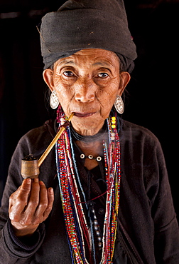 Woman of the Ann tribe in traditional black dress and colourful beads smoking a pipe in the doorway of her home in a hill village near Kengtung (Kyaingtong), Shan State, Myanmar (Burma), Asia