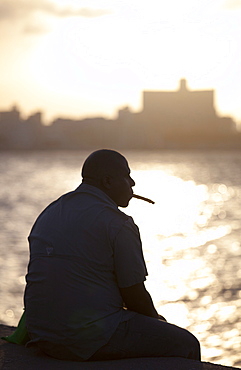 Man in semi silhouette sat on the sea wall at sunset, smoking a cigar, The Malecon, Havana Centro, Havana, Cuba, West Indies, Central America