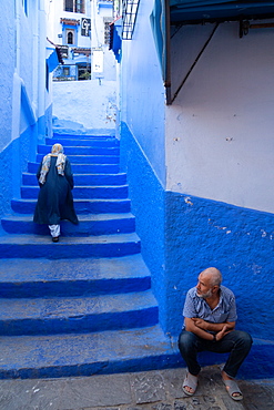 Woman in dark blue traditional clothes climbing up the stairs of alleyway, man seated in the foreground, Chefchaouen, Morocco, North Africa, Africa