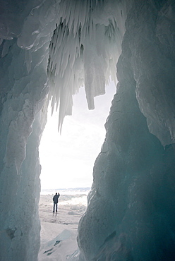 Tourist framed in the frozen mouth of ice cave, with icicles formed on roof of caves at Olkhon Island as the waves freeze in winter, Lake Baikal, Irkutsk Oblast, Siberia, Russia, Eurasia