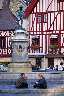 A couple sit in front of a fountain in the Place Francois Rude, with half timbered houses behind, in Dijon, Burgundy, France, Europe