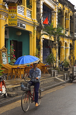 Man riding a bike and holding umbrella, passing typical colonial buildings in the old town, Hoi An, Vietnam, Indochina, Southeast Asia, Asia