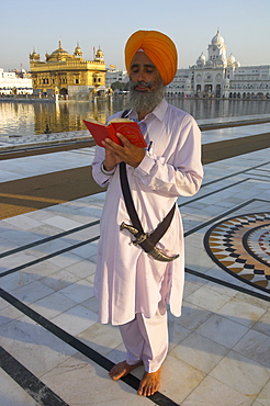 Sikh pilgrim with orange turban, white dress and dragger, reading prayer book, in front of the Golden Temple, Amritsar, Punjab state, India, Asia