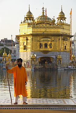 Shrine guard in orange clothes holding lance standing by pool in front of the Golden Temple, Amritsar, Punjab, India, Asia