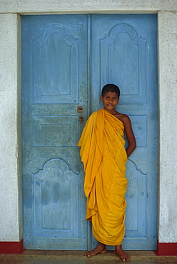 Portrait of a young Buddhist monk in saffron robe in front of a blue door near Kandy, Sri Lanka, Asia