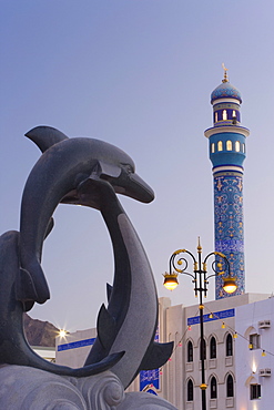 Mosque minaret and dolphin sculpture on the Mutrah Corniche illuminated at dusk, Mutrah, Muscat, Oman, Middle East