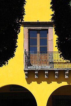 Mexico, Bajio, San Miguel de Allende, El Jardin detail of yellow painted exterior facade of colonial mansion with French windows and balcony part framed by silhouetted trees.