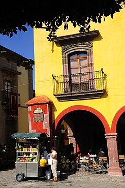 Mexico, Bajio, San Miguel de Allende, El Jardin Part view of yellow painted facade of colonial mansion with French window and balcony with fruit juice vendor in street below.