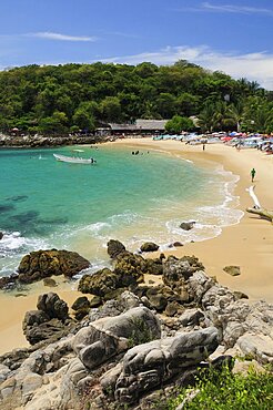 Mexico, Oaxaca, Puerto Escondido, View onto Playa Manzanillo beach with rocks in foreground and tourist boats people and tree covered headland beyond.